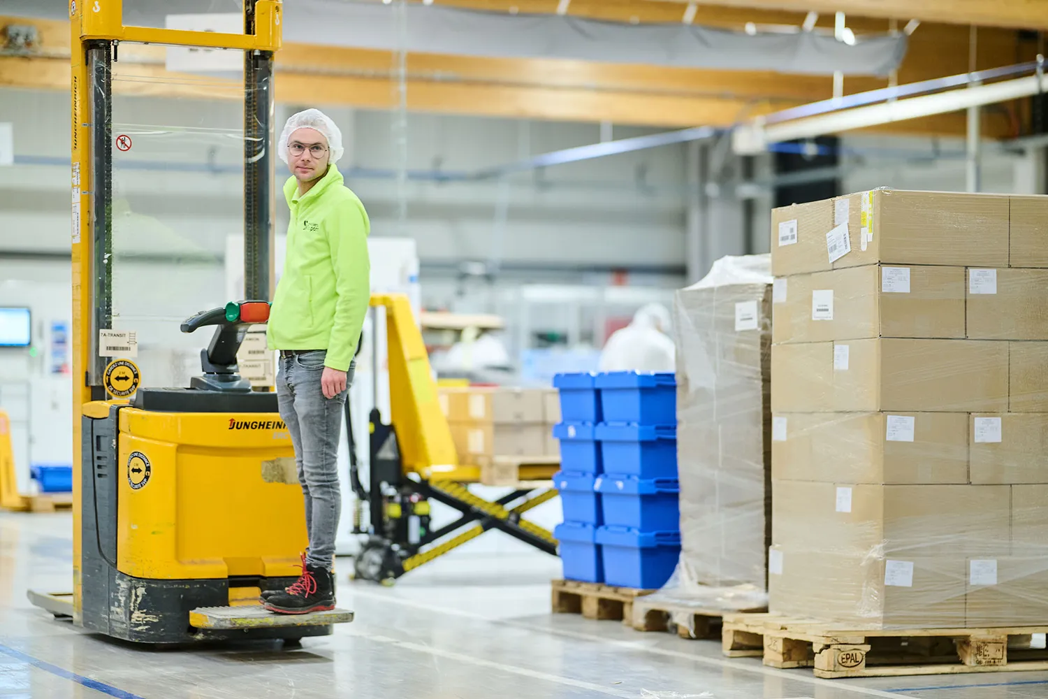 CONPALUX-81150 Un homme en gilet vert fluo se tient sur un transpalette jaune dans un entrepôt avec des palettes de cartons et de bacs bleus.