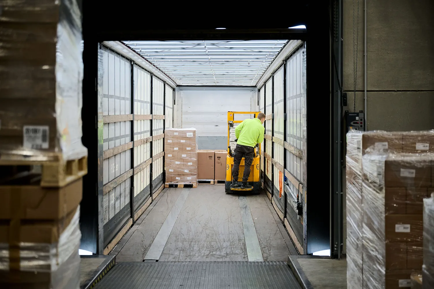 CONPALUX-82586 Un homme en gilet vert fluo utilise un transpalette dans un camion, entouré de palettes de cartons emballés.