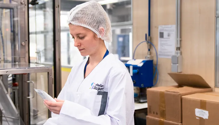 CONPALUX-chaine Une personne en blouse blanche et charlotte lit un document dans une usine avec des cartons et machines en arrière-plan.