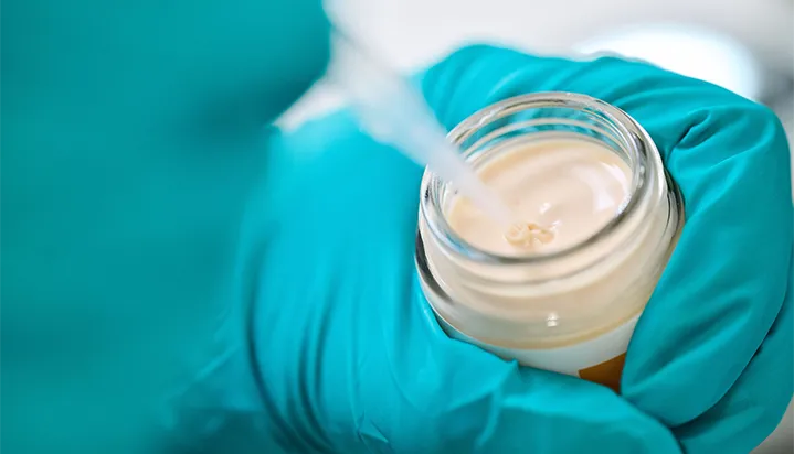CONPALUX-production Des mains gantées de bleu tiennent un pot ouvert de crème beige, avec une pipette insérée dans le produit.